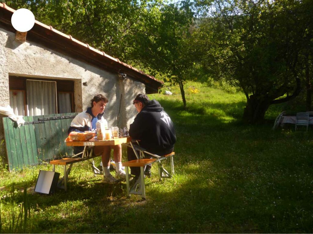 Amis partageant un repas face à face devant une maison de campagne, autour d’une table pliable installée sur l’herbe ensoleillée.