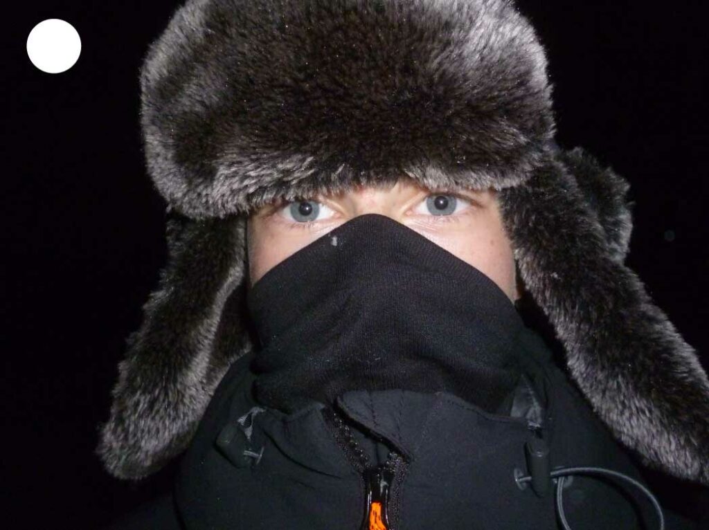 Portrait nocturne d’un garçon aux yeux bleus éclairé au flash, portant une chapka, avec une mise au point précise sur le regard pour un effet percutant.