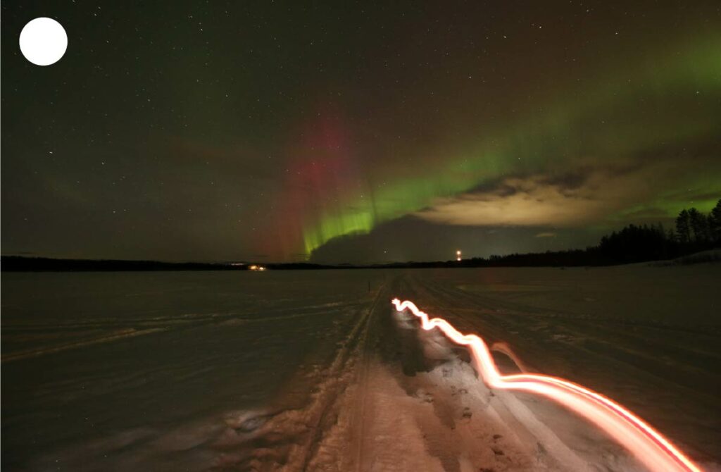 Aurore boréale verte et rouge traversant le ciel nocturne en Laponie, avec une ligne lumineuse se reflétant sur la neige blanche.